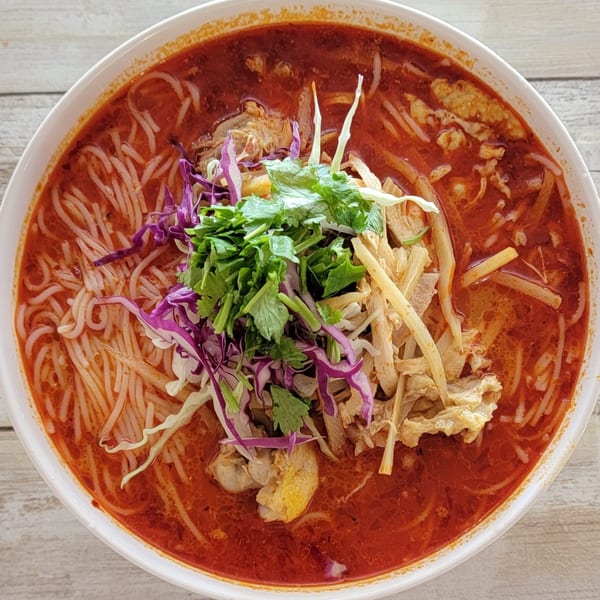 A vibrant bowl of Som Jai Thai Cuisine's red curry noodle soup, garnished with fresh herbs and cabbage.
