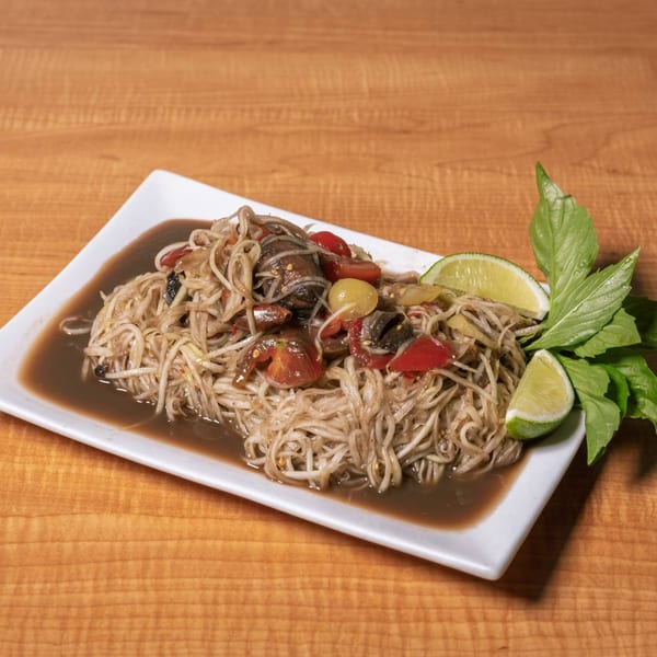 Som Tum, a Thai papaya salad with noodles, tomatoes, and lime wedges on a white plate.