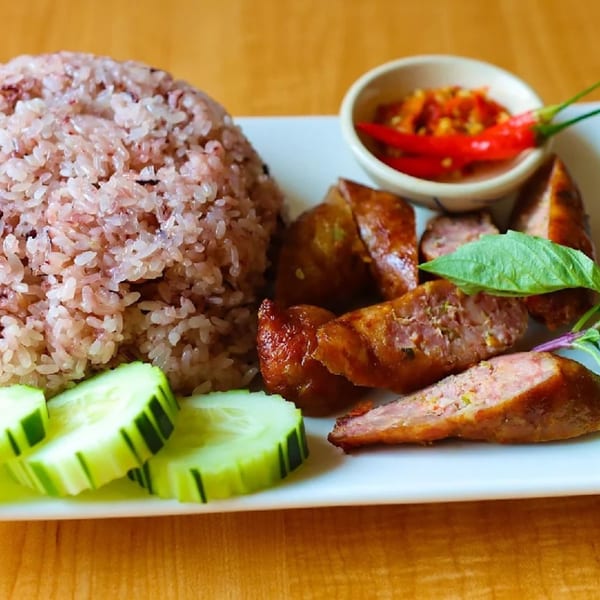 Thai sausage with red rice, sliced cucumbers, and chili sauce on a white plate.